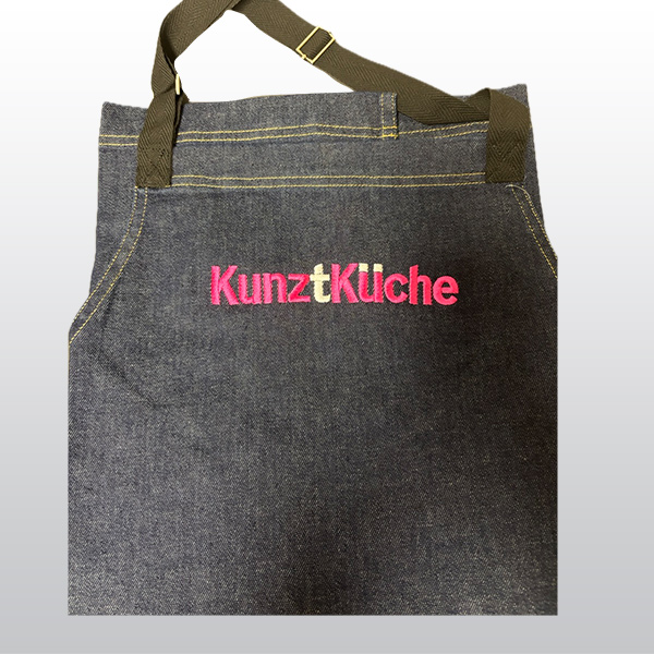 Eine knielange Schürze aus dunklem Jeansstopp. Die Hinz&Kunzt Stickerei "KunztKüche" ist in magentafarben und weiß auf Brusthöhe aufgestickt.