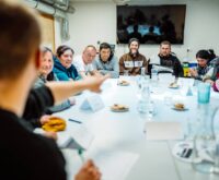 Eine Gruppe von Menschen sitzt an einem Tisch, alle schauen zum Mann am Tischkopf