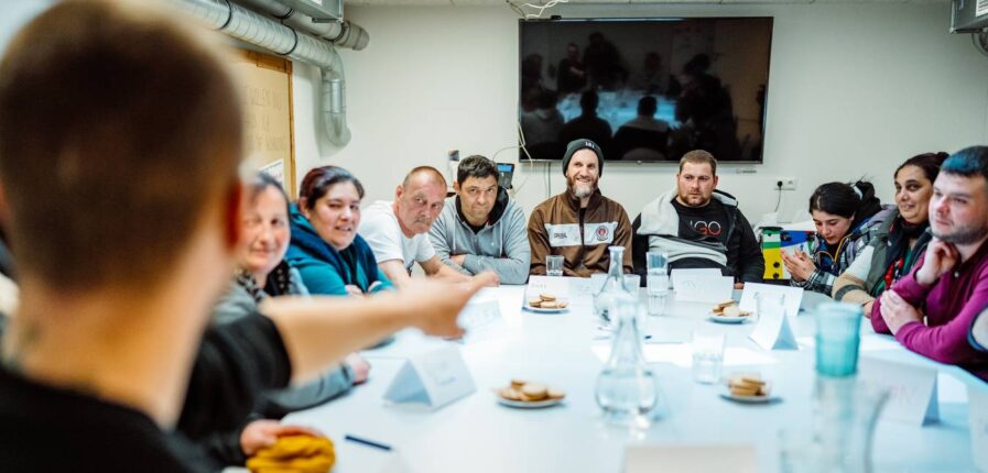 Eine Gruppe von Menschen sitzt an einem Tisch, alle schauen zum Mann am Tischkopf