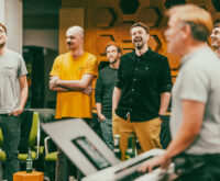 Vier Männer stehen in einem Raum und singen, einer spielt Keyboard