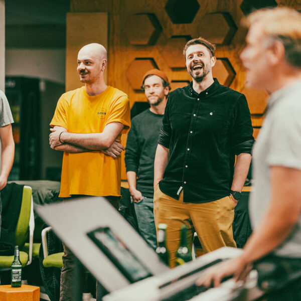 Vier Männer stehen in einem Raum und singen, einer spielt Keyboard