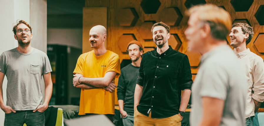 Vier Männer stehen in einem Raum und singen, einer spielt Keyboard