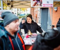 Ein Mann mit Mütze steht hinter einem Tresen vor einem Backsteinhaus