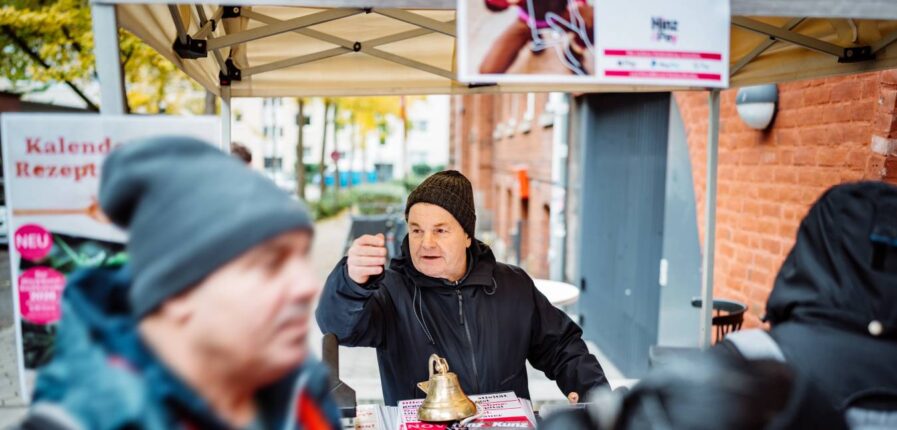 Ein Mann mit Mütze steht hinter einem Tresen vor einem Backsteinhaus