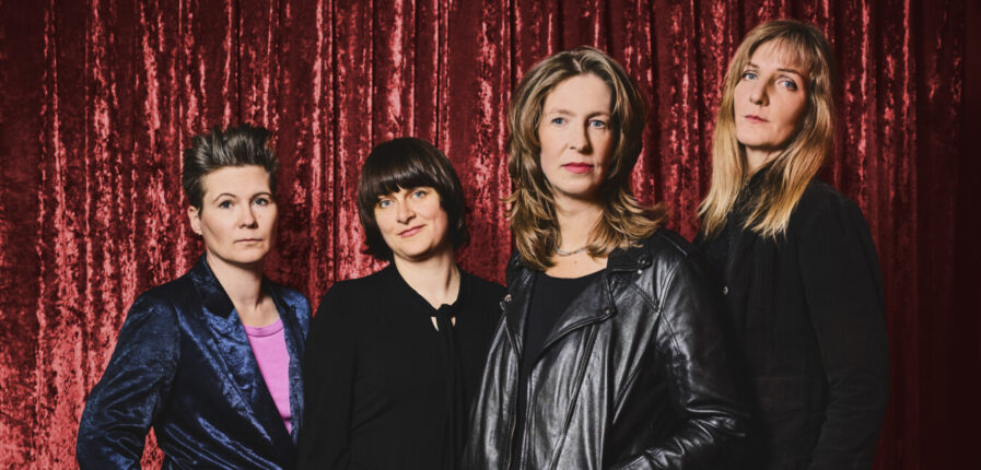 Vier Frauen stehen vor einem roten Vorhang und schauen ernst in die Kamera