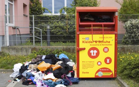Ein Altkleidercontainer. Daneben liegt ein Haufen Klamotten auf der Erde.