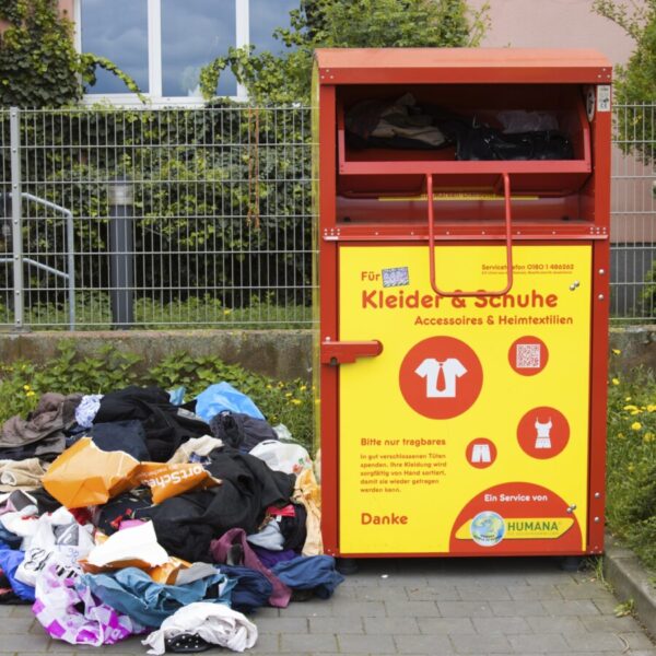 Ein Altkleidercontainer. Daneben liegt ein Haufen Klamotten auf der Erde.