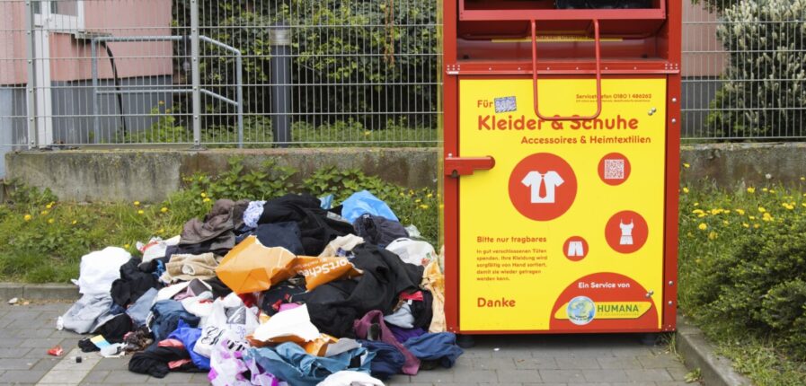 Ein Altkleidercontainer. Daneben liegt ein Haufen Klamotten auf der Erde.