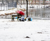 Ein obdachloser Mensch sitzt auf einer Bank, neben ihm sein Hab und Gut. Der Boden ist mit Schnee bedeckt. Im Hintergrund ist ein zugefrorener Teich zu sehen.