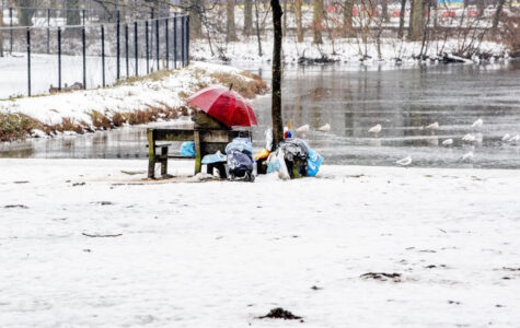 Ein obdachloser Mensch sitzt auf einer Bank, neben ihm sein Hab und Gut. Der Boden ist mit Schnee bedeckt. Im Hintergrund ist ein zugefrorener Teich zu sehen.