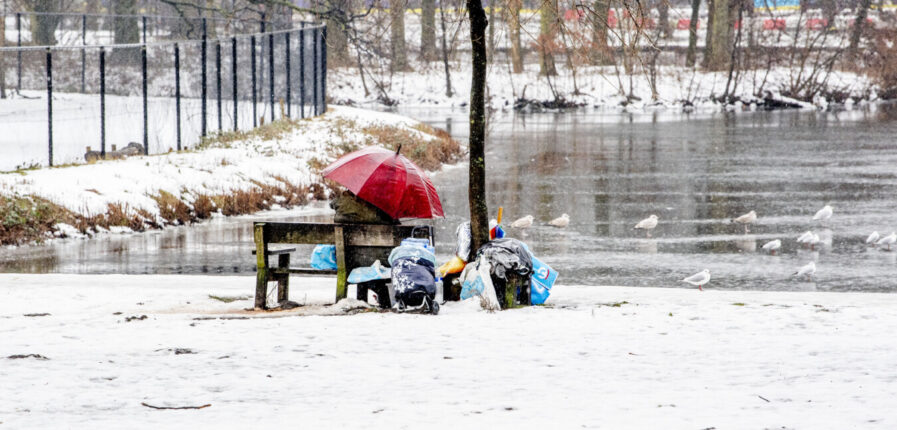 Ein obdachloser Mensch sitzt auf einer Bank, neben ihm sein Hab und Gut. Der Boden ist mit Schnee bedeckt. Im Hintergrund ist ein zugefrorener Teich zu sehen.