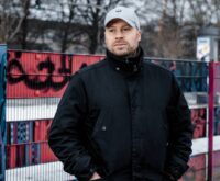 Ein Mann mit grauer Kappe und dunkler steht vor einem bunten Zaun