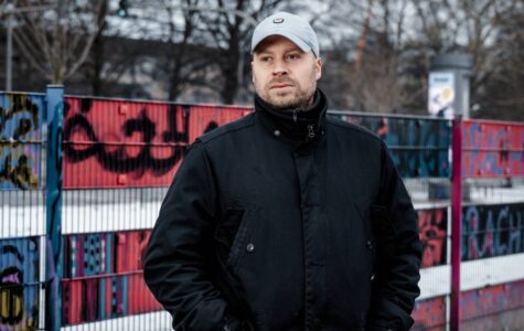 Ein Mann mit grauer Kappe und dunkler steht vor einem bunten Zaun