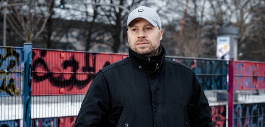 Ein Mann mit grauer Kappe und dunkler steht vor einem bunten Zaun