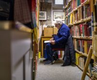 Ein grauhaariger Mann sitzt auf einem Stuhl zwischen bücherregalen