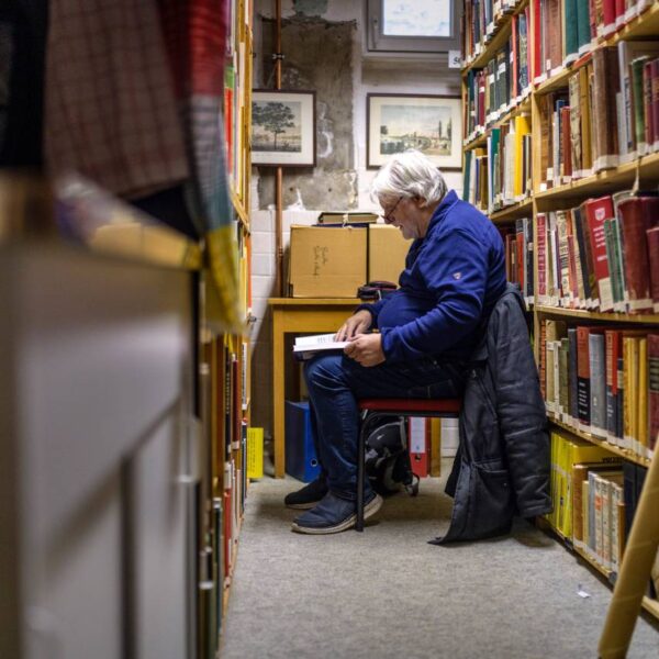 Ein grauhaariger Mann sitzt auf einem Stuhl zwischen bücherregalen