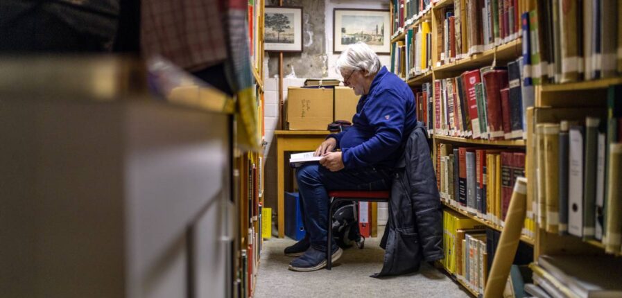 Ein grauhaariger Mann sitzt auf einem Stuhl zwischen bücherregalen