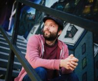 Ein Mann mit Vollbart, Mütze und im Hoodie sitzt auf einer Treppe