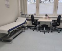 Ein Behandlungszimmer mit zwei Stühlen, einem Tisch und einer Krankenliege.