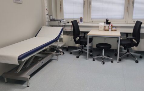 Ein Behandlungszimmer mit zwei Stühlen, einem Tisch und einer Krankenliege.