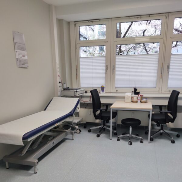 Ein Behandlungszimmer mit zwei Stühlen, einem Tisch und einer Krankenliege.