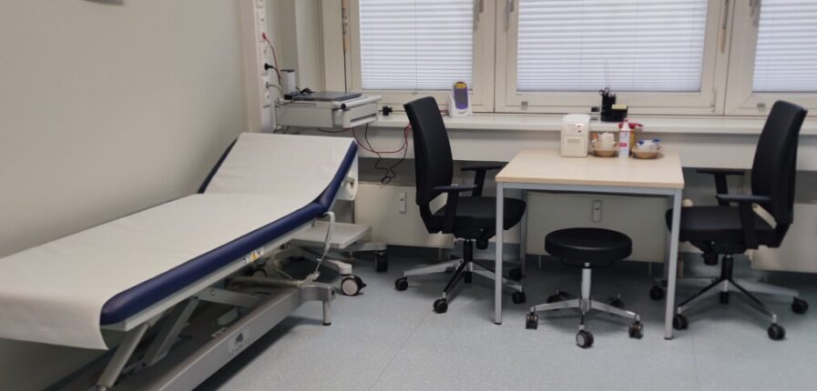 Ein Behandlungszimmer mit zwei Stühlen, einem Tisch und einer Krankenliege.