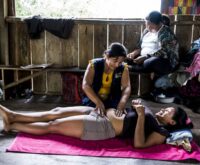 Eine schwangere Frau liegt rücklings auf dem Boden, eine andere kniet neben ihr und tastet den Bauch ab