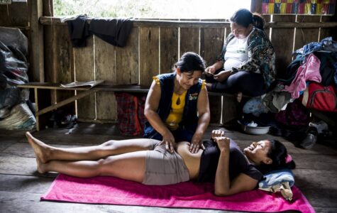 Eine schwangere Frau liegt rücklings auf dem Boden, eine andere kniet neben ihr und tastet den Bauch ab
