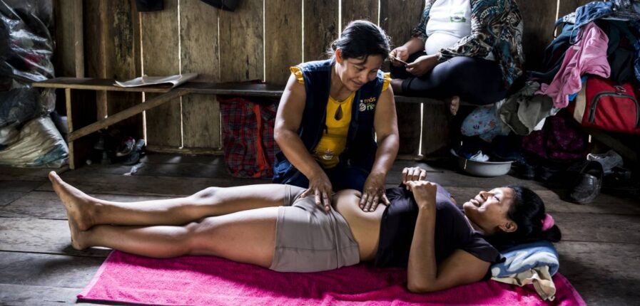 Eine schwangere Frau liegt rücklings auf dem Boden, eine andere kniet neben ihr und tastet den Bauch ab