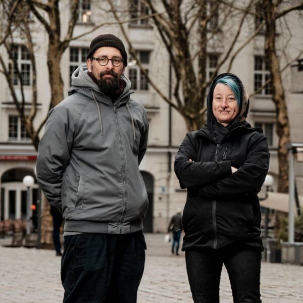 Ein Mann mit Mütze und eine Frau mit grünen Haaren schauen in die Kamera