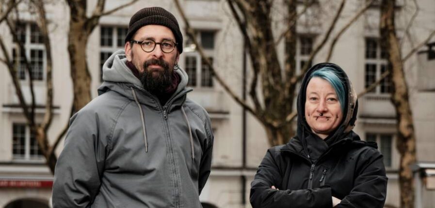 Ein Mann mit Mütze und eine Frau mit grünen Haaren schauen in die Kamera