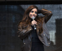 Eine Frau mit braunen Haaren und brauner Lederjacke hält ein Mikrofon in der Hand