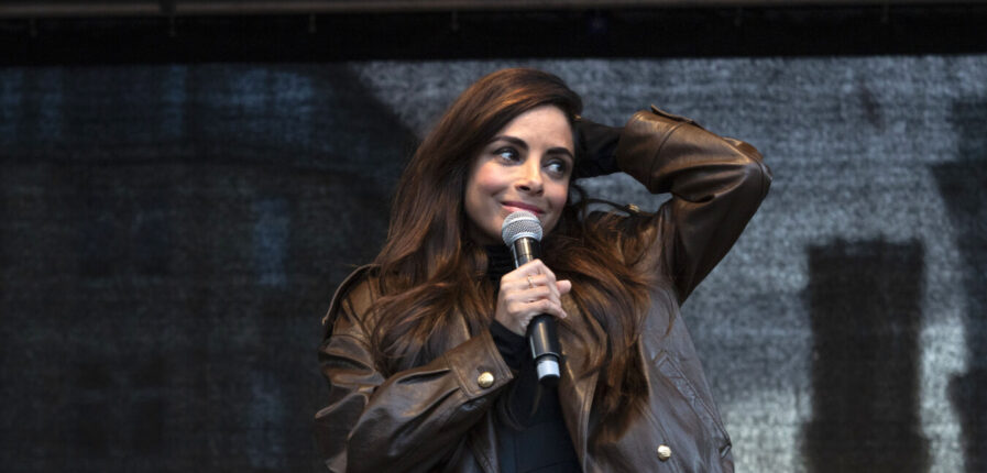 Eine Frau mit braunen Haaren und brauner Lederjacke hält ein Mikrofon in der Hand