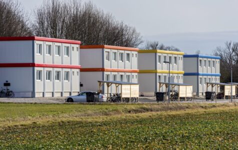 Drei Gebäude aus gestapelten grau-blauen Containern, davor Fahrräder und ein Auto