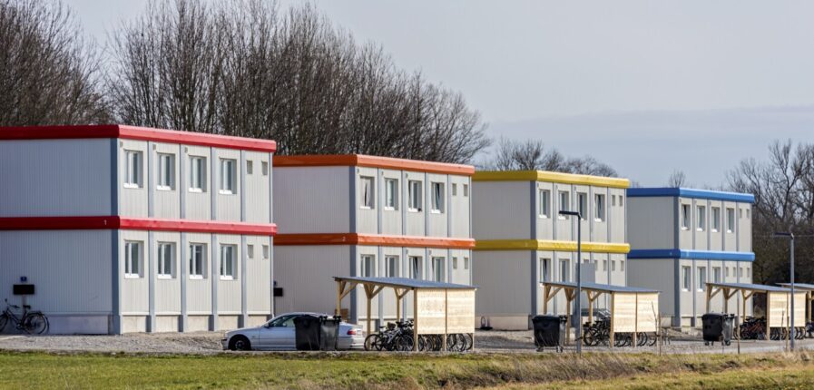 Drei Gebäude aus gestapelten grau-blauen Containern, davor Fahrräder und ein Auto