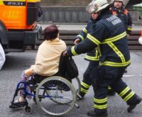 Eine Frau im Rollstuhl wird von zwei Feuerwehrleuten geschoben