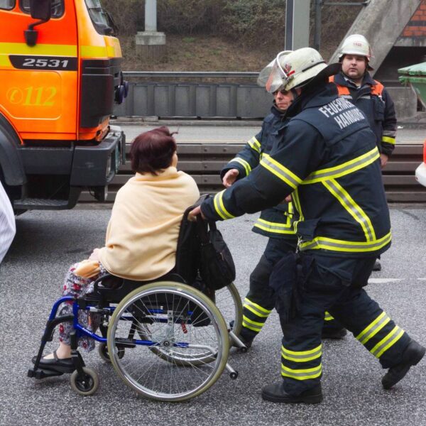 Eine Frau im Rollstuhl wird von zwei Feuerwehrleuten geschoben