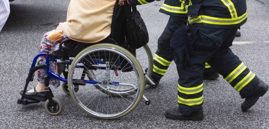 Eine Frau im Rollstuhl wird von zwei Feuerwehrleuten geschoben