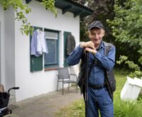 Ein Mann steht vor einem kleinen Haus und schaut freundlich in die Kamera