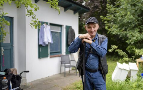 Ein Mann steht vor einem kleinen Haus und schaut freundlich in die Kamera