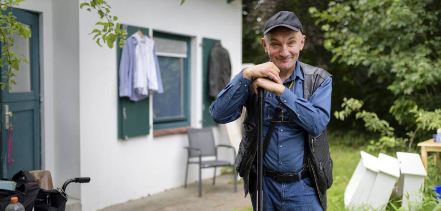 Ein Mann steht vor einem kleinen Haus und schaut freundlich in die Kamera