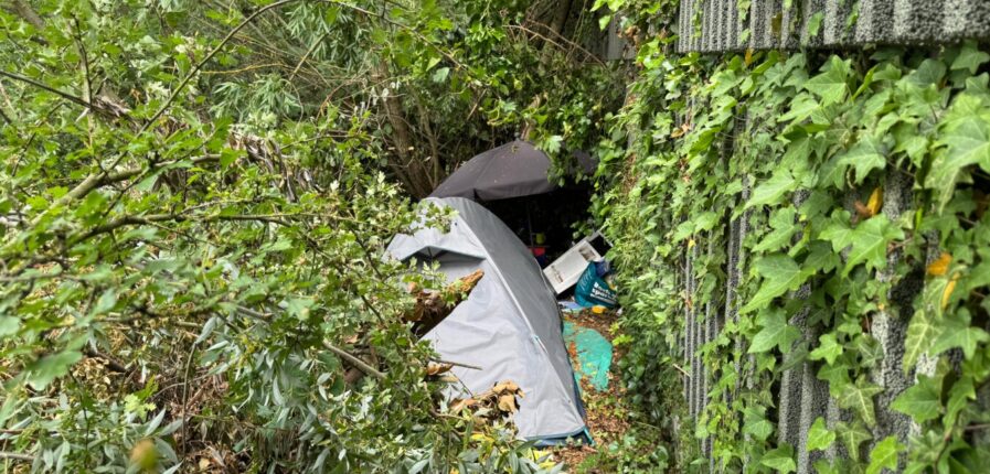 Rechts: Eine graue Lärmschutzwand. Direkt daran anschließend steht ein Zelt im Gebüsch.