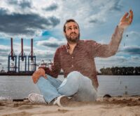 Ein Mann sitzt am Elbstrand und lässt Sand durch seine Finger laufen. Im Hintergrund sieht man Hafenkräne.
