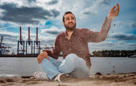 Ein Mann sitzt am Elbstrand und lässt Sand durch seine Finger laufen. Im Hintergrund sieht man Hafenkräne.