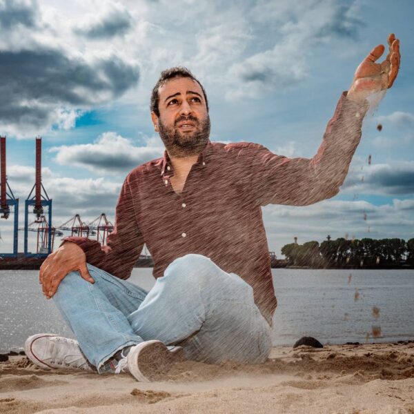 Ein Mann sitzt am Elbstrand und lässt Sand durch seine Finger laufen. Im Hintergrund sieht man Hafenkräne.