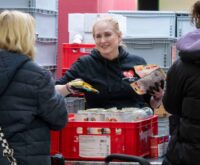 Eine blonde Frau gibt Tierfutter an zwei Menschen aus, die man von hinten sieht