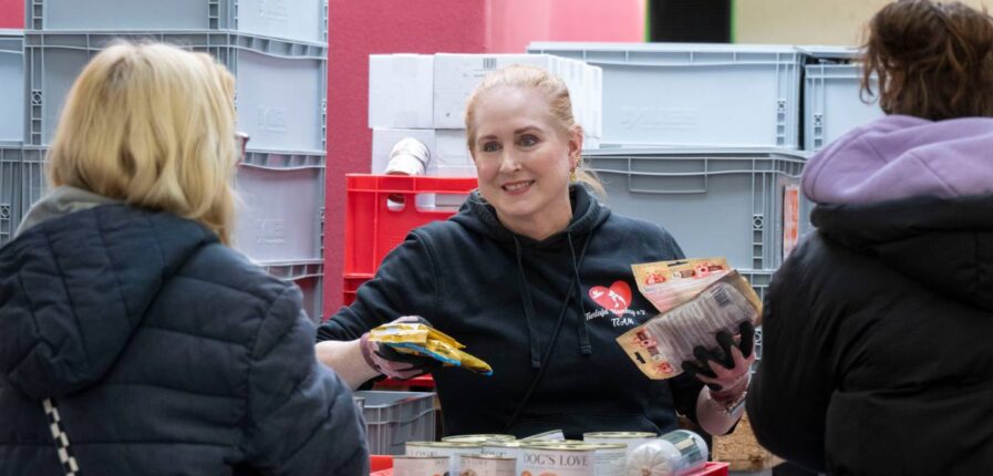 Eine blonde Frau gibt Tierfutter an zwei Menschen aus, die man von hinten sieht