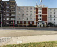 Rechts ist ein grauses Haus mit vielen Fenstern zu sehen. Auf dem Dach steht in großen Buchstaben: Hotel. Links daneben: Ein Wohnhaus.