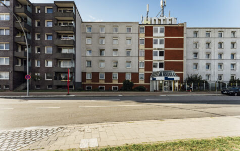 Rechts ist ein grauses Haus mit vielen Fenstern zu sehen. Auf dem Dach steht in großen Buchstaben: Hotel. Links daneben: Ein Wohnhaus.