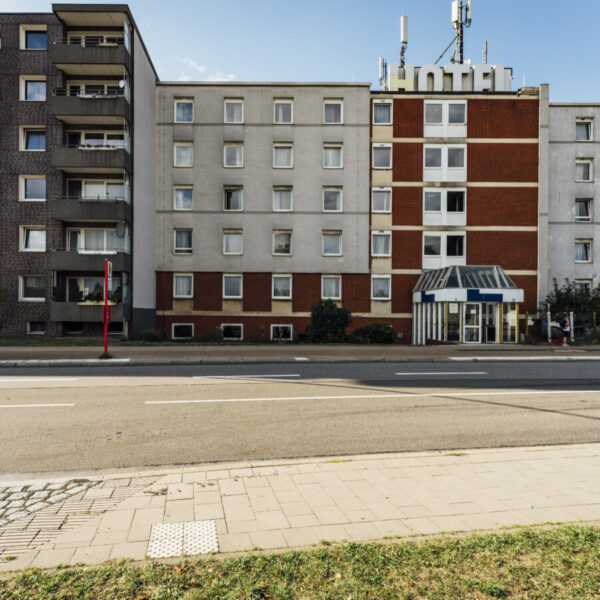 Rechts ist ein grauses Haus mit vielen Fenstern zu sehen. Auf dem Dach steht in großen Buchstaben: Hotel. Links daneben: Ein Wohnhaus.